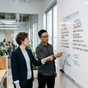 Two professionals standing by a whiteboard, discussing AI strategy options and diagrams in an office setting.