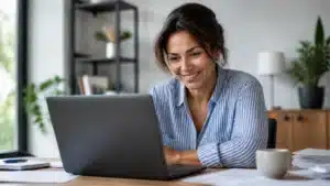 Woman smiling while working on a laptop at a tidy home office desk with papers and a mug nearby